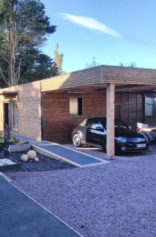 Maison moderne en bois avec une voiture garée sous un auvent et un aménagement paysager.